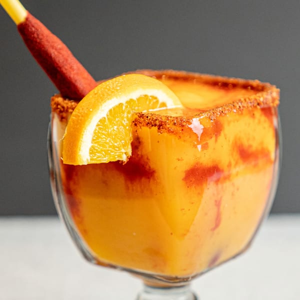 A vibrant orange Mexican drink in a glass, garnished with an orange slice and a red candy stick.