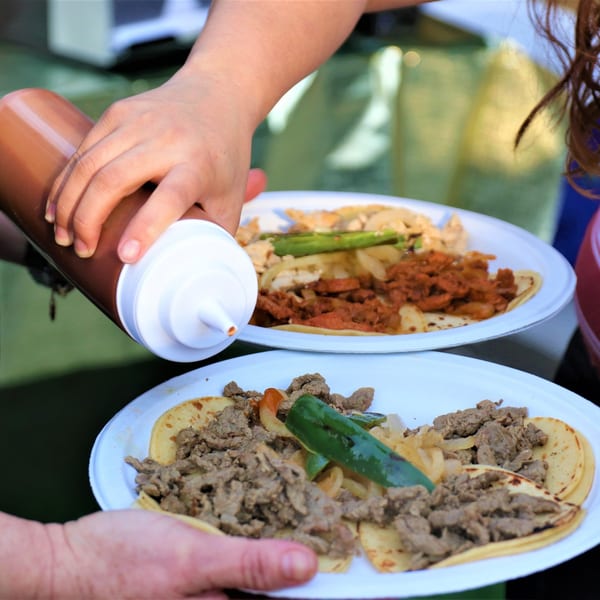 Hands squeezing sauce onto two plates of El Paso Mexican Restaurant tacos with various fillings.