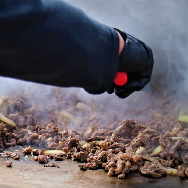 Chef in black gloves cooking sizzling fajita meat and onions on a flat top grill.
