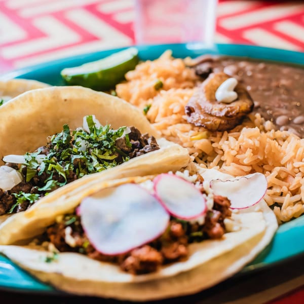 Two tacos with meat, cilantro, and radishes, served with rice, beans, and lime on a blue plate.