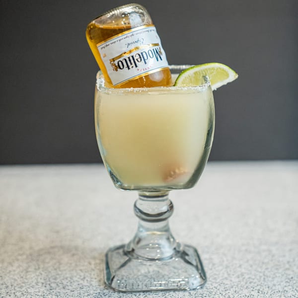 A Modelo beer bottle inverted into a margarita glass with a salted rim and lime wedge.