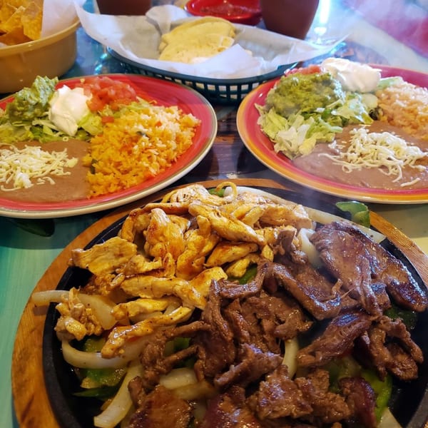 Sizzling chicken and beef fajitas with onions and peppers, alongside plates of rice, beans, and guacamole.