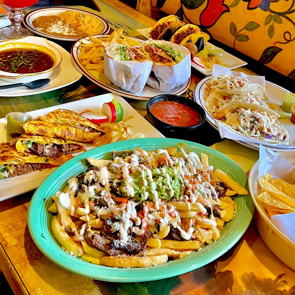 A table laden with various Mexican dishes, including tacos, burritos, quesadillas, and loaded fries.