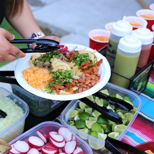A person holds a plate of tacos with rice and beans, surrounded by toppings and sauces.