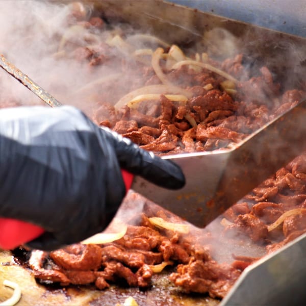 A gloved hand uses a spatula to stir sizzling El Paso Mexican Restaurant fajita meat and onions on a grill.